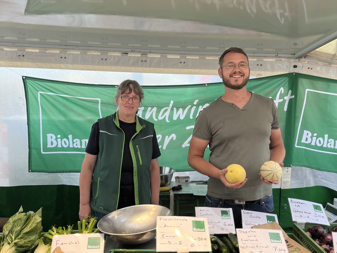 Zwei Verkäufer hinter einem Wochenmarktstand mit Biolebensmitteln
