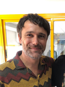 KI generiert: Das Bild zeigt einen lächelnden Mann mit Bart in einem farbig gemusterten Hemd. Im Hintergrund ist ein gelber Rahmen und ein lichtdurchfluteter Raum zu sehen.