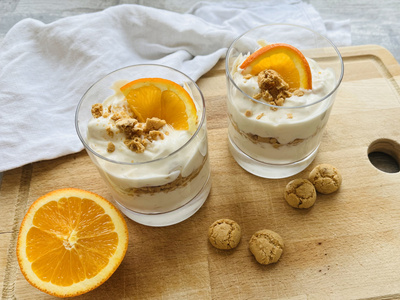 Dessert im Glas mit Sahne, Kekskrümeln und Orangenscheiben, auf Holzbrett serviert.