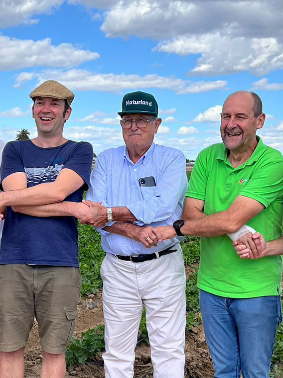 KI generiert: Drei Männer stehen lächelnd auf einem Feld und halten einander die Hände. Einer der Männer trägt eine Kappe mit der Aufschrift "Naturland".