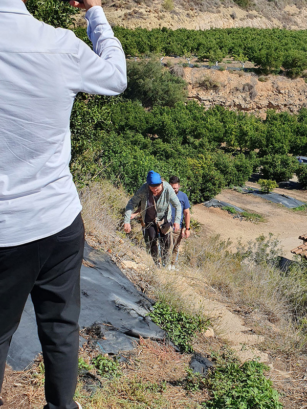 KI generiert: Das Bild zeigt drei Personen, die einen Hang in einer ländlichen Gegend hinaufsteigen. Im Hintergrund sind üppige grüne Pflanzungen und terrassierte Hügel zu sehen.