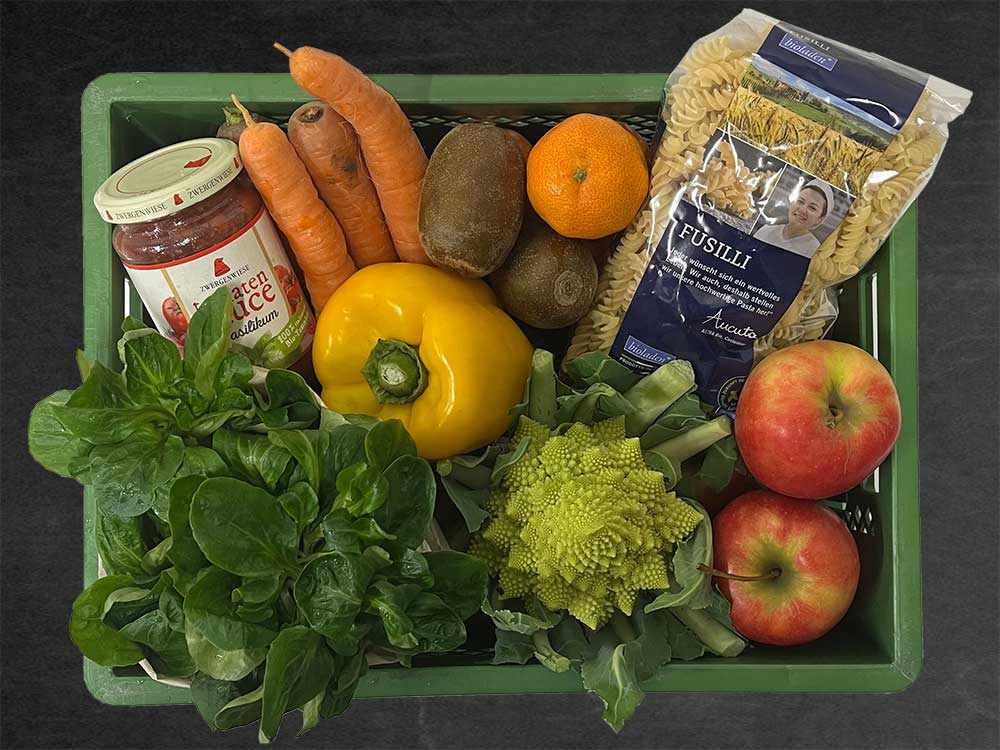 KI generiert: Das Bild zeigt eine grüne Kiste, die verschiedene Lebensmittel enthält, darunter frisches Gemüse wie Romanesco, Karotten, eine gelbe Paprika, Obst wie Äpfel und Kiwis, sowie Nudeln und Tomatensauce. Es handelt sich um eine Auswahl an frischen und verpackten Lebensmitteln.