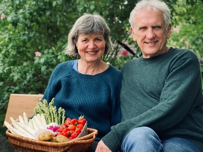 Zwei lächelnde Personen halten einen Korb voller frischem Gemüse und Obst im Freien.