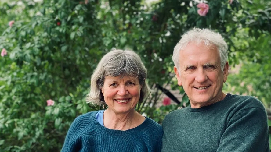 KI generiert: Ein älteres Paar lächelt in einem Garten mit grünen Büschen und rosafarbenen Blumen.