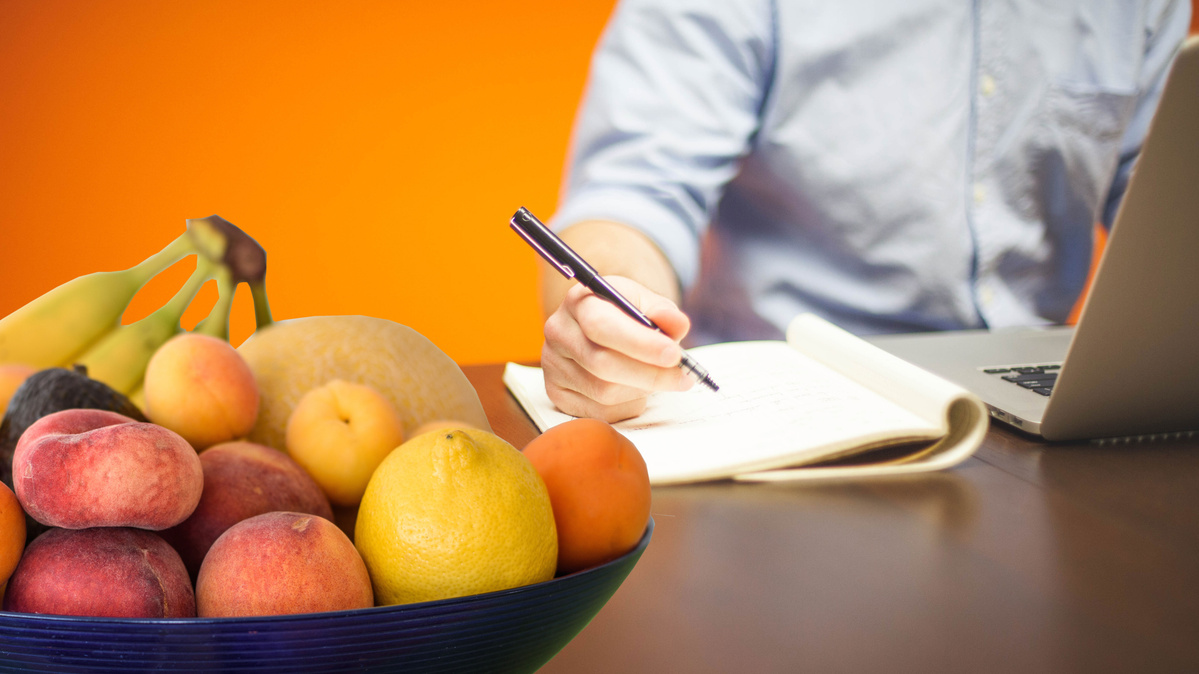 Jemand sitzt am Schreibtisch vor einem Laptop und macht sich Notizen. Im Vordergrund befindet sich eine Schale mit Obst
