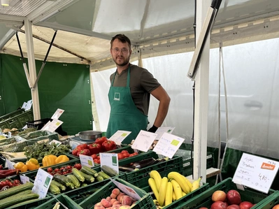 Marktstand mit Mann in grüner Schürze, der frisches Obst und Gemüse verkauft. Text: "Äpfel Braeburn 4,80."