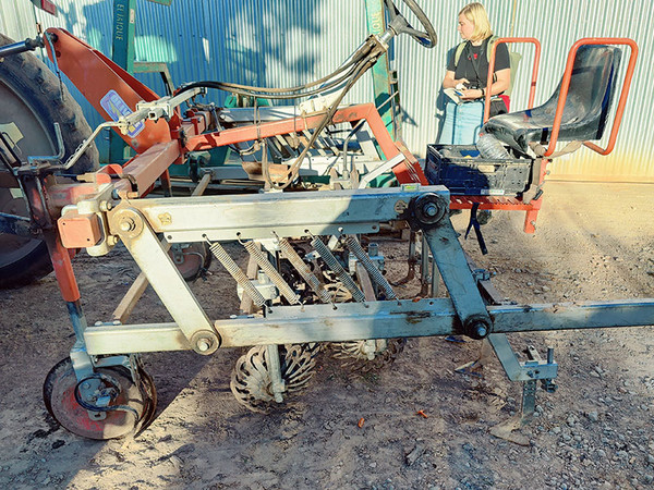 KI generiert: Das Bild zeigt eine landwirtschaftliche Maschine mit einem Sattelplatz und mechanischen Komponenten im Vordergrund, während eine Person im Hintergrund steht. Es scheint sich um eine Art Erntemaschine oder Sämaschine zu handeln, die neben eines Traktors positioniert ist.