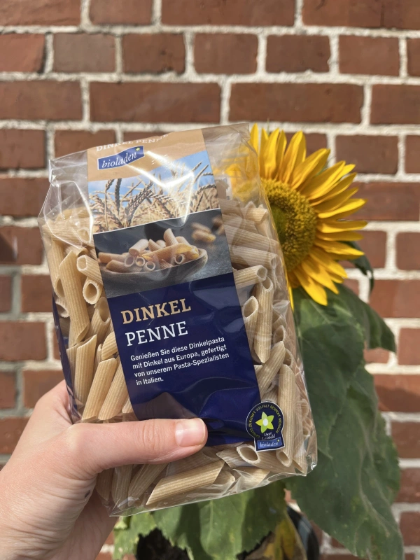 KI generiert: Packung Dinkel-Penne vor Sonnenblume und Backsteinmauer. Text: "DINKEL PENNE"