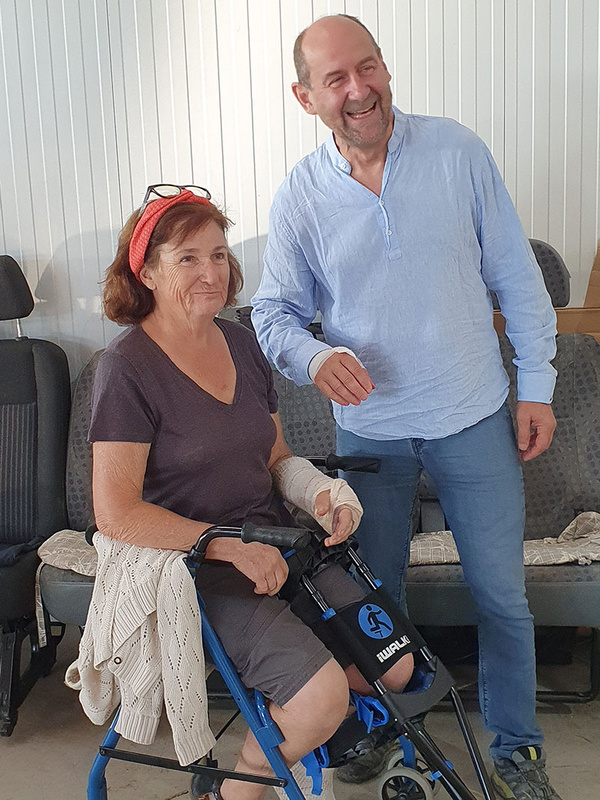 KI generiert: Eine Frau sitzt lächelnd auf einem Rollator, während ein Mann neben ihr steht und ebenfalls lächelt. Beide befinden sich in einem Raum mit Autositzen im Hintergrund.