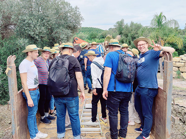 KI generiert: Eine Gruppe von Menschen mit Hüten und Rucksäcken steht auf einer Holzplattform in einer ländlichen Umgebung. Einer der Personen zeigt ein Peace-Zeichen und lächelt in die Kamera.