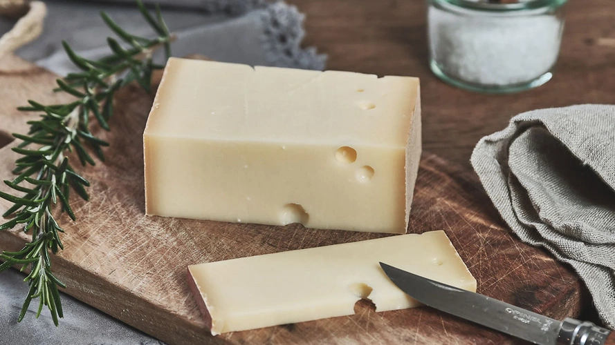 Ein Stück Käse mit Löchern auf einem Holzbrett, daneben ein Messer und Rosmarinzweig.
