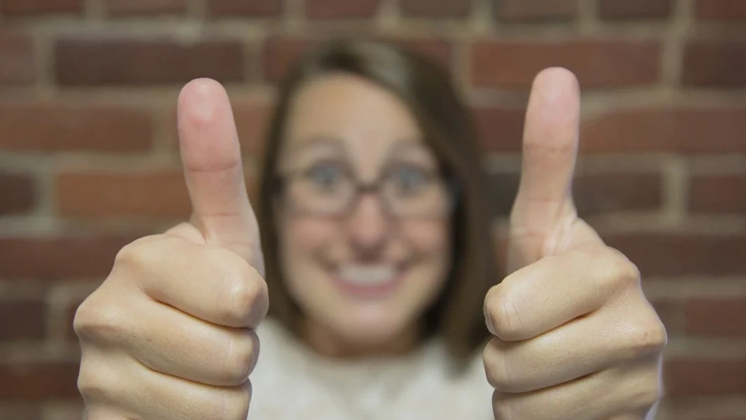 Eine Frau steht vor einer Ziegelwand und hält beide Daumen nach oben. Die Hände sind im Focus. Das Gesicht ist unscharf