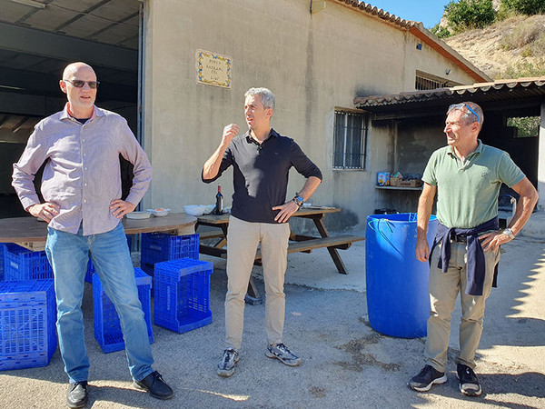 KI generiert: Drei Männer stehen entspannt vor einem Gebäude im Freien und unterhalten sich. Neben ihnen sind blaue Kisten und ein Tisch mit Flaschen und Schalen zu sehen.