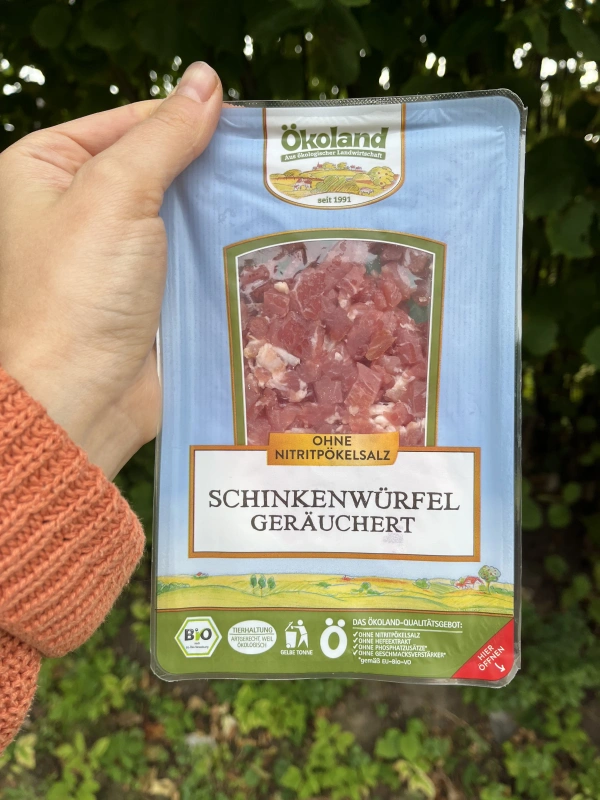 KI generiert: Eine Hand hält eine "Ökoland" Packung mit geräucherten Schinkenwürfeln. Text: "OHNE NITRITPÖKELSALZ."