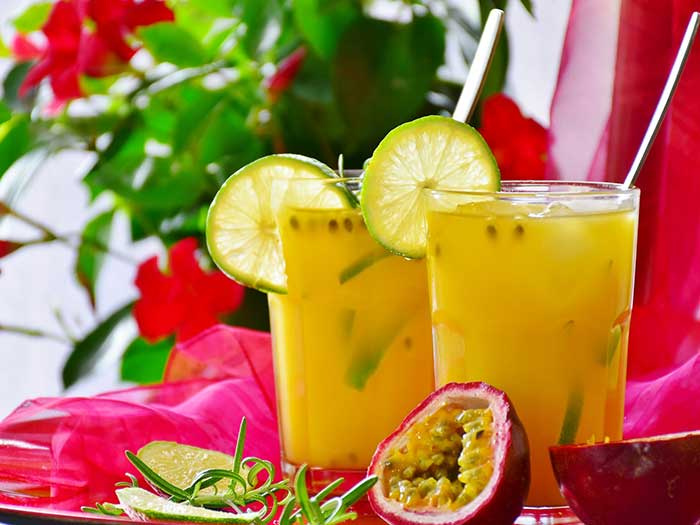 Zwei Gläser, gefüllt mit Fruchtsaft und mit Zironenscheiben am Glasrand dekoriert stehen auf rotem Tuch. Im Vordergrund eine durchgeschnittene Marakuya Frucht. Im Hintergrund eine Unscharfe Pflanze.