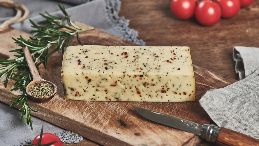 Ein Stück Käse mit Kräutern auf einem Holzbrett, daneben Rosmarin und Tomaten.