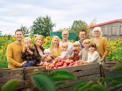 Eine Familie steht lächelnd vor Kisten voller Äpfel auf einem Sonnenblumenfeld.