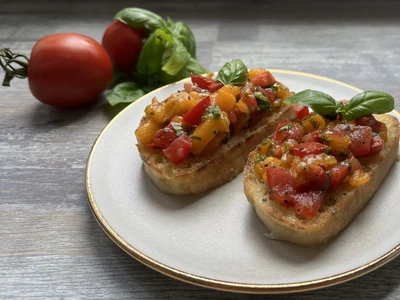 Zwei Bruschette mit Tomaten-Mix und Basilikum auf einem Teller, daneben frische Tomaten und Basilikum.