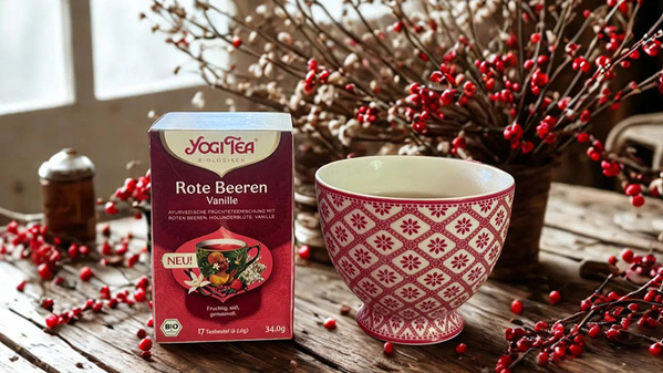 Teepackung "Yogi Tea Rote Beeren Vanille" und eine Tasse mit Beeren im Hintergrund auf einem Holztisch.