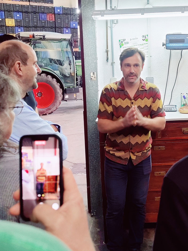 KI generiert: Eine Person in einem gemusterten Hemd hält einen Vortrag oder eine Präsentation in einer Halle, möglicherweise einer Lager- oder Produktionsstätte, während ein anderer Mensch das Geschehen mit einem Smartphone filmt. Im Hintergrund ist ein Traktor und gestapelte Kisten zu sehen.