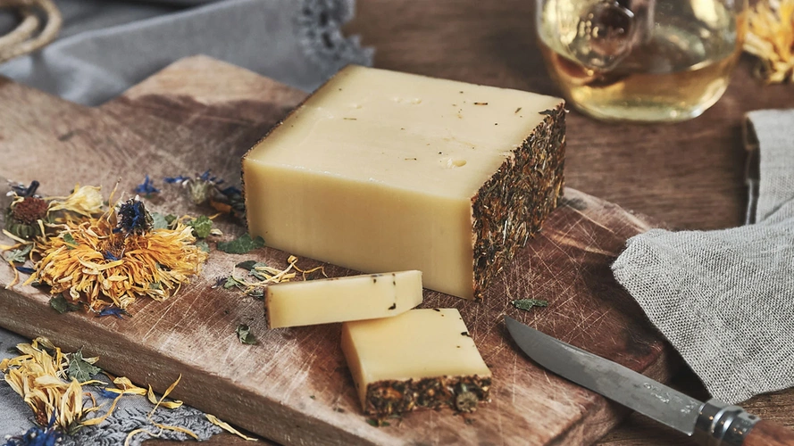 Käseblock mit Kräuterrand auf Holzbrett, daneben geschnittene Stücke und ein Messer. Deko: getrocknete Blüten.