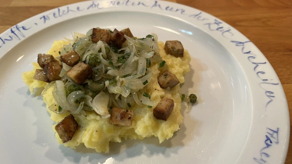 KI generiert: Auf einem Teller: Kartoffelbrei mit Zwiebeln und Tofuwürfeln. Text: „Liebe, welche die Zeiten durchquert.“