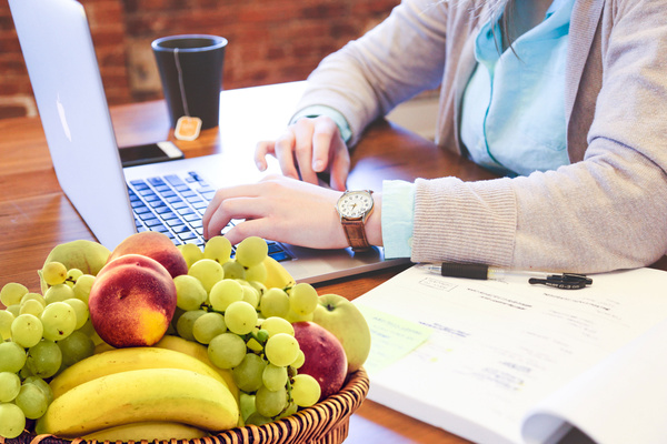 Jemand sitzt am Schreibtisch vor einem Laptop. Im Vordergrund befindet sich eine Schale mit Obst