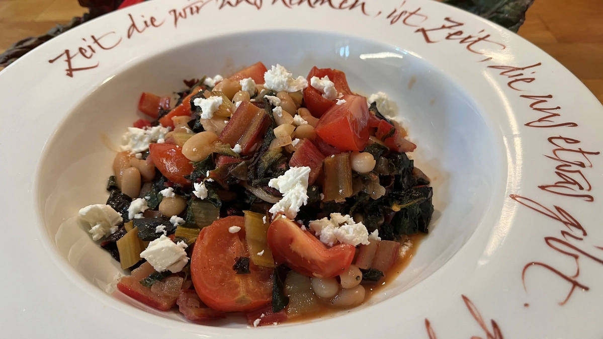 Ein Teller mit buntem Mangold, weißen Bohnen, Tomaten und Feta. Der Text auf dem Tellerrand lautet: "Zeit, die wir uns nehmen, ist Zeit, die uns etwas gibt."