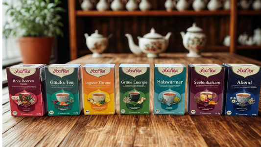 Verschiedene Yogi Tea-Packungen auf einem Tisch mit Hintergrundeinrichtung aus Teekannen. Text: "Rote Beeren Vanille, Glücks Tee, Ingwer Zitrone, Grüne Energie, Halswärmer, Seelenbalsam, Abend."