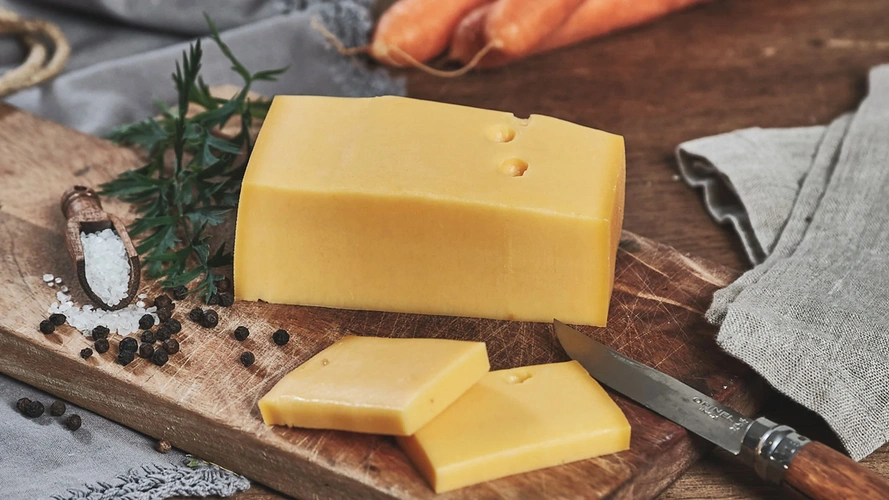 Ein Stück gelber Käse mit zwei Scheiben und einem Messer auf einem Holzbrett, umgeben von Salz, Pfeffer und Möhren.