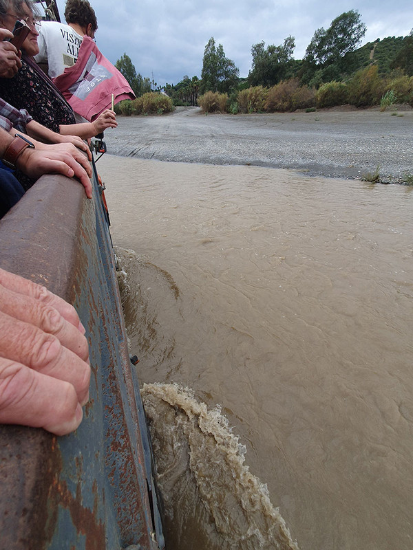 KI generiert: Das Bild zeigt Menschen, die in einem Fahrzeug sitzen, das einen schlammigen Fluss überquert. Im Hintergrund sind Bäume und ein bewölkter Himmel zu sehen.