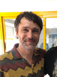 KI generiert: Das Bild zeigt einen lächelnden Mann mit Bart in einem farbig gemusterten Hemd. Im Hintergrund ist ein gelber Rahmen und ein lichtdurchfluteter Raum zu sehen.