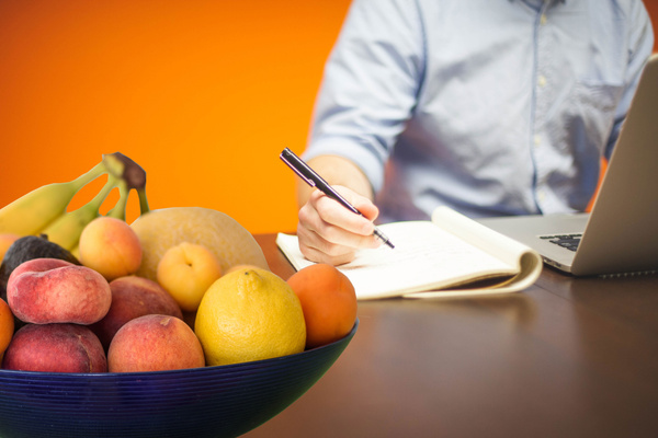 Jemand sitzt am Schreibtisch vor einem Laptop und macht sich Notizen. Im Vordergrund befindet sich eine Schale mit Obst