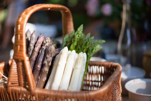 KI generiert: Ein Korb mit grünem, weißem und violettem Spargel, im Hintergrund unscharf.