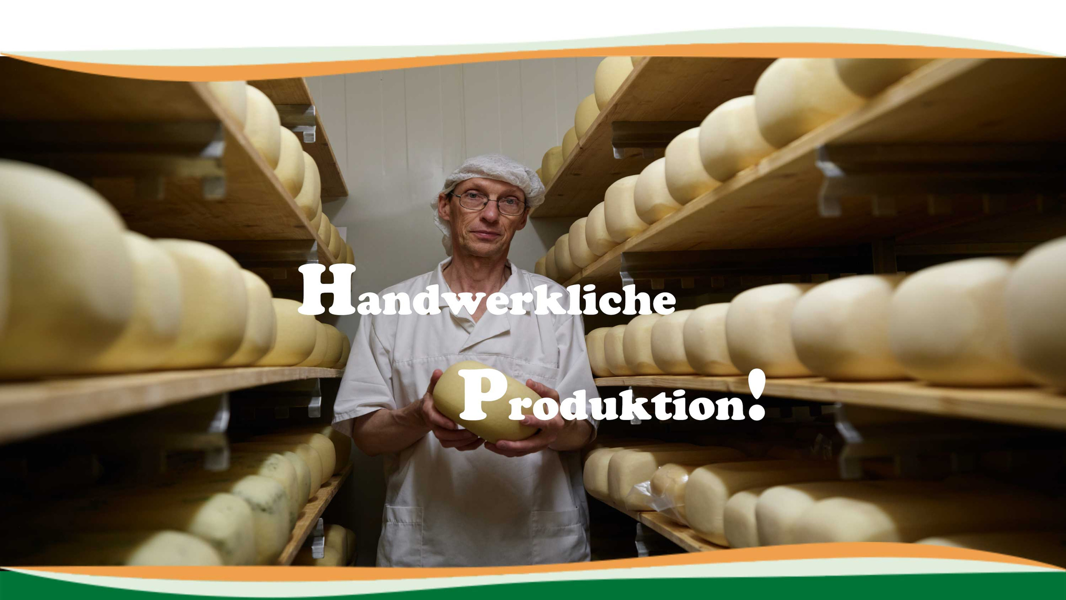 Ein Käser zeigt einen ganzen Käse. Er steht in einem Käse-Reiferaum. Beidseitig liegen Käse in mehreren Etagen auf Holzregalen.